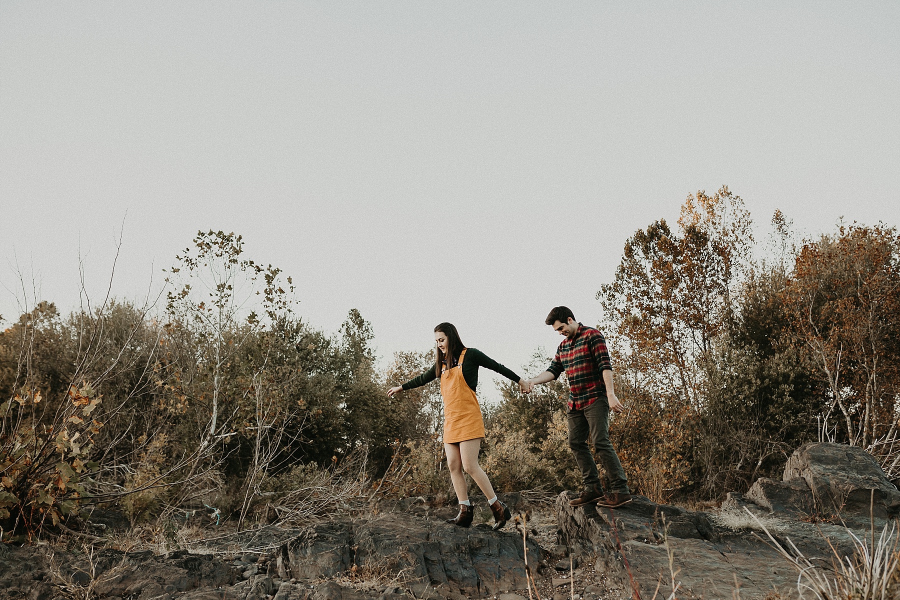 Couple holding hands as they walk along the top of a rocky ledge as the sun sets in front of them